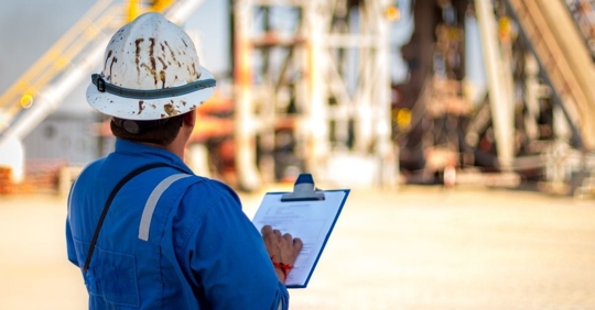 man wearing a blue jumpsuit and dirt-covered hard-hat holding a clipboard and looking at an oil rig